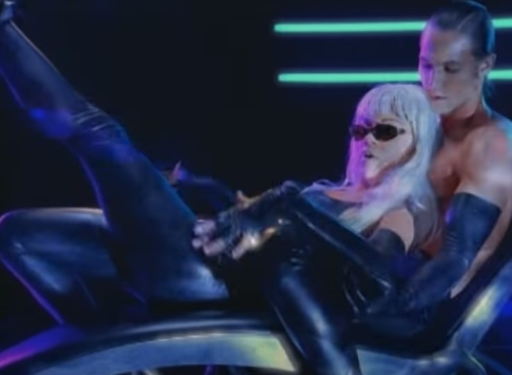 A couple in skintight black catsuits in front of neon lights on a dark background.
